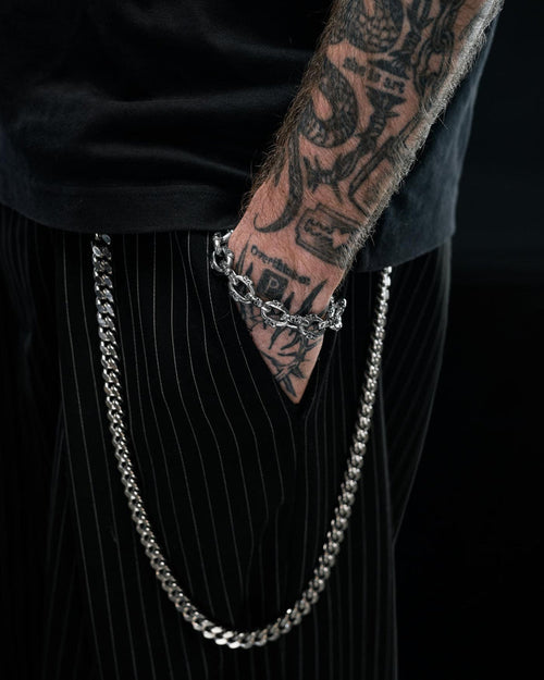 A tattooed arm rests by black pinstripe pants with a silver chain hanging from the waistband, wearing the Statement Collective "Boneyards" Skull Bracelet on the wrist, set against a dark background.