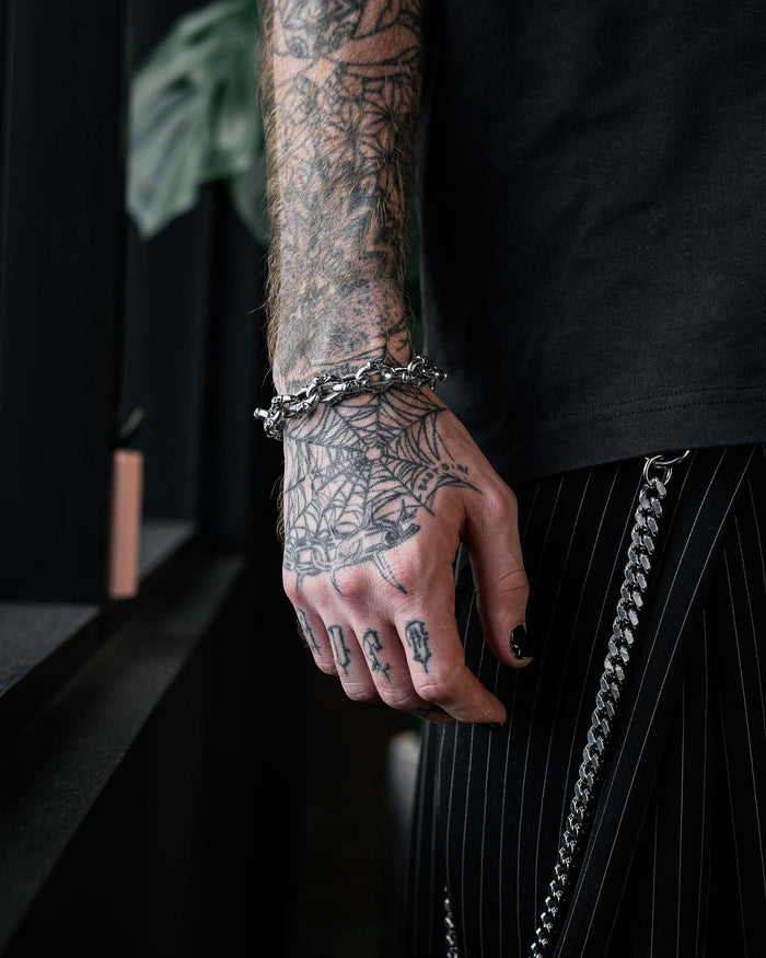 A tattooed hand adorned with the "Boneyards" Skull Bracelet by Statement Collective and rings rests by the side, wearing black pinstripe pants with chain accessories. The person sports black nail polish and a web tattoo on their hand.