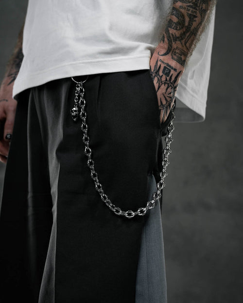 A person wearing black and gray pants with the "Boneyards" Skull Wallet Chain clipped to the pocket stands with a tattooed hand in the pocket, a partially visible white shirt, and a dark, blurred background.
