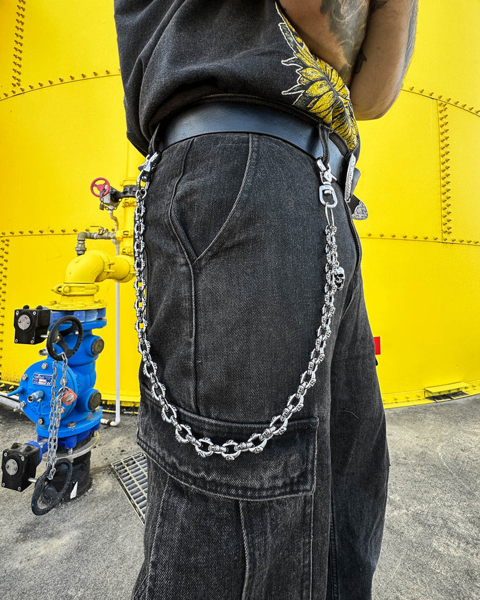 A person wears black pants with the "Boneyards" Skull Wallet Chain clipped to a belt loop, standing by a bright yellow industrial tank and blue fire hydrant. Only their lower torso and hip are visible.