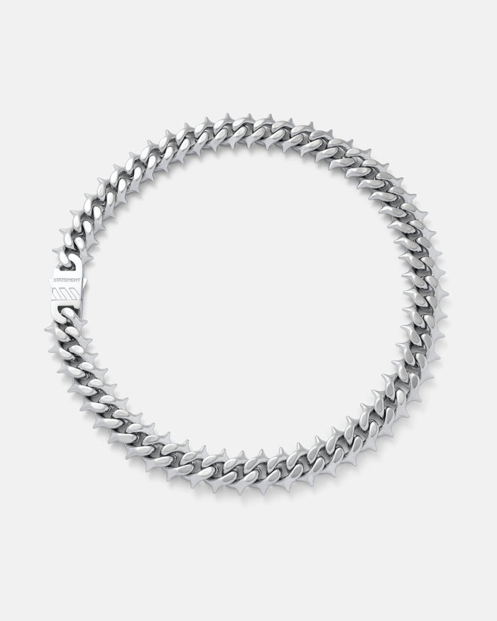 The "Chainsaw" Spiked Cuban Chain, a silver stainless steel necklace with interlocking links and clasp, is displayed in a circular shape on a plain light gray background.