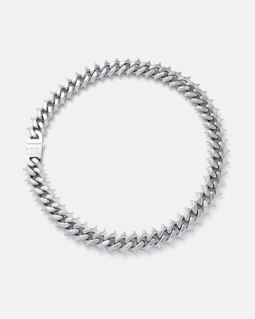 The "Chainsaw" Spiked Cuban Chain, a silver stainless steel necklace with interlocking links and clasp, is displayed in a circular shape on a plain light gray background.