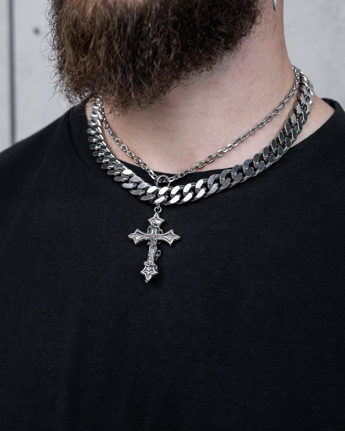 A person with a partial beard wears a black shirt and Statement Collective's Chunky Ruins Set—a layered necklace set featuring silver chains, including a Cuban link and a stainless steel detailed cross pendant.