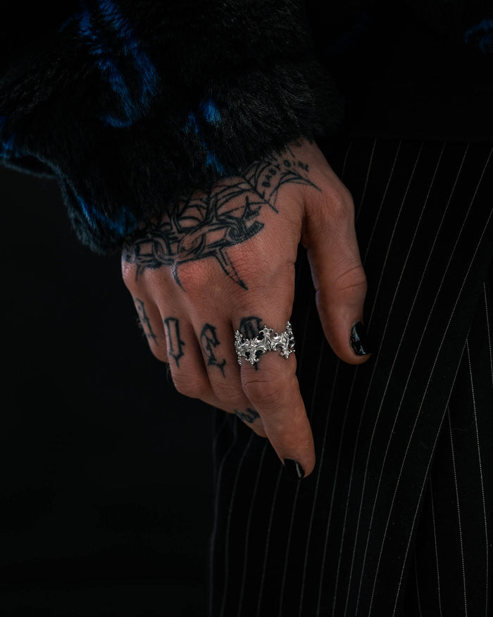 A tattooed hand with black-painted nails displays a detailed silver bat-shaped ring alongside the eye-catching “Crested” Fleur De Lis Ring by Statement Collective. The wearer’s dark fuzzy sleeve and pinstriped pants stand out against a black background, enhancing the bold aesthetic.