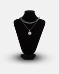 A black necklace display stand showcases the Crucifix Set: one chunky barbed wire necklace and one slim chain with a circular fallen angel medallion pendant.