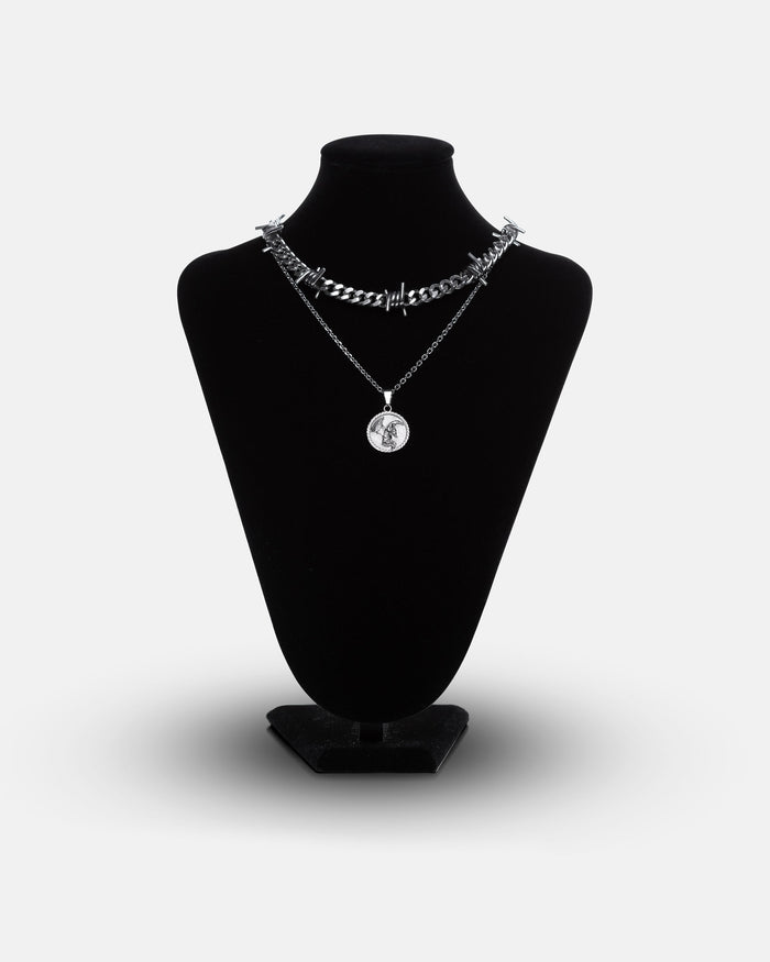 A black necklace display stand showcases the Crucifix Set: one chunky barbed wire necklace and one slim chain with a circular fallen angel medallion pendant.