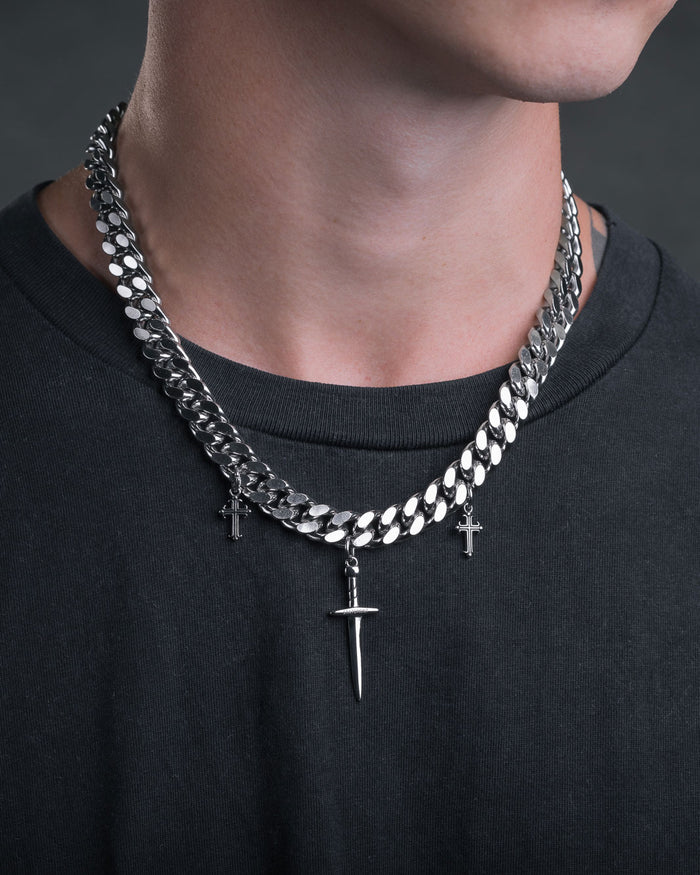 Wearing a black shirt, the person sports two silver chain necklaces: a punk-style one with a large spike pendant and the "Crusader" 11mm Cuban Chain featuring two small cross pendants, all set against a dark background.