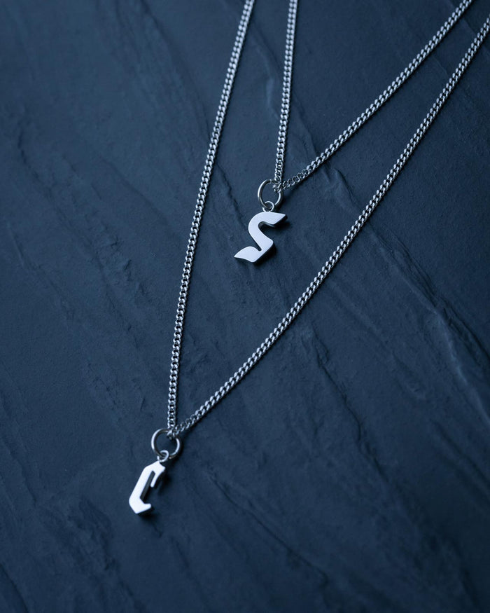 Two silver curb chains with Custom Initial Letter Pendants—S and C—from Statement Collective rest on a dark stone surface, perfect for fans of personalized necklaces.
