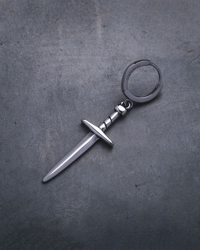 The Dagger Earring—a silver accessory shaped like a small dagger with a straight blade and crossguard, reminiscent of statement jewelry—rests on a textured gray surface.