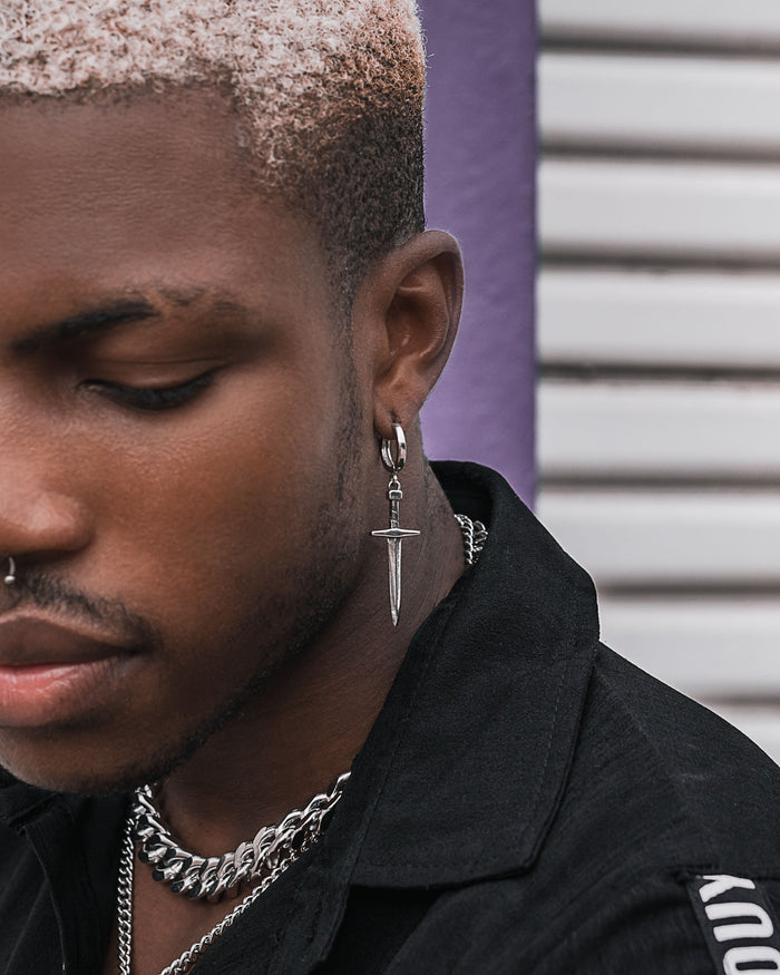 A person with short, bleached hair wears the Dagger Earring, a silver nose ring, and layered chain necklaces as statement jewelry with a black shirt, standing before a striped background.