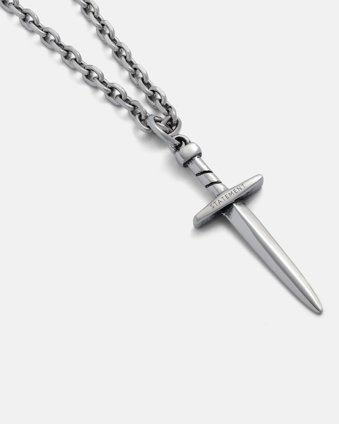 The Dagger Pendant is a silver chain necklace with an engraved "STATEMENT" hilt, shown on a light gray background—ideal for fans of hand-carved jewelry.