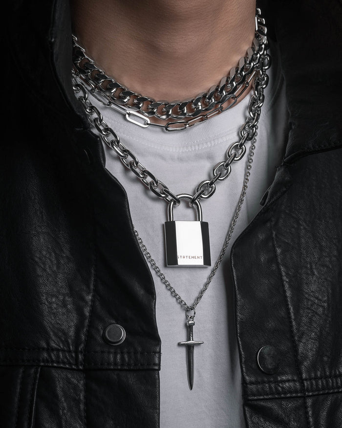A person wearing the Statement Collective Handle With Care 2.0 Set—a stylish men's necklace stack featuring layered silver necklaces: a chunky chain link with padlock pendant, a bar pendant chain, and a thin sword pendant—over a white shirt and black leather jacket for the perfect modern statement jewelry look.