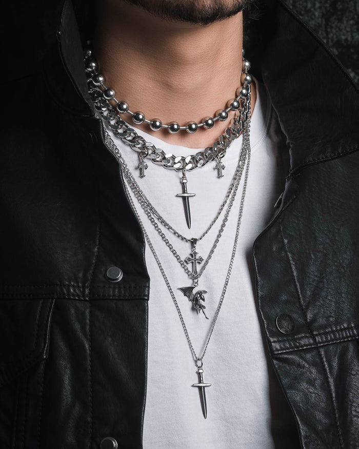 A person wearing the Statement Collective Sharp Objects Set—layered silver necklaces featuring a bold cuban link chain along with cross and dagger pendants—styled over a white shirt and black leather jacket. The image highlights the striking jewelry and fashionable outfit, with the face out of focus.