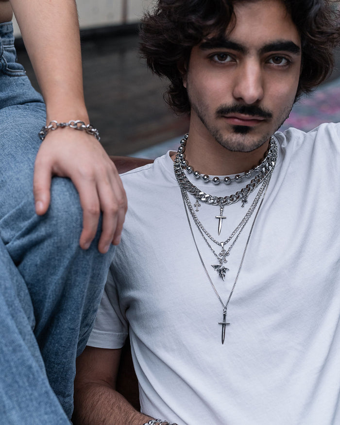 A person with wavy hair and a mustache wears layered silver necklaces from the Statement Collective Sharp Objects Set, including a bold statement piece, paired with a white t-shirt. They are seated next to another individual in blue jeans whose arm and hand are visible.