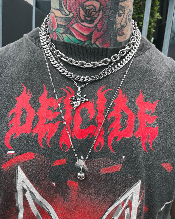 A person wearing the Dead Love Set by Statement Collective—a grunge necklace set featuring layered silver chains and a bold skull heart pendant—over a black shirt emblazoned with “DEICIDE” in red flame lettering. The individual also has vibrant neck tattoos and some visible face tattoos.