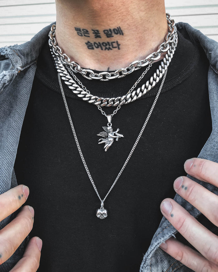 A person wearing the Dead Love Set by Statement Collective, a grunge necklace set with layered silver chains featuring fairy and skull heart pendants. They have a Korean tattoo on their neck, small tattoos on their fingers, and are styled in a partially unbuttoned denim jacket.