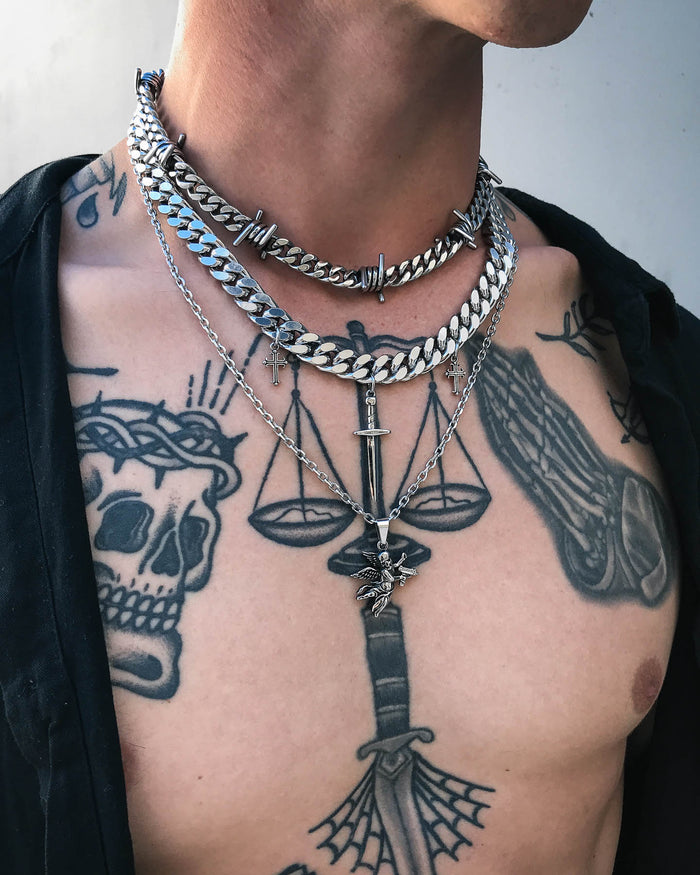 Wearing a black shirt open to display tattoos of a skull, scales of justice, a sword, and webbing, this person layers multiple silver chains and necklaces featuring barbed wire designs and the Death's Cupid Pendant.