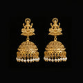 A pair of gold-toned Indian jhumka earrings with detailed filigree work, small white pearl bead drops, and deity motifs at the top, displayed on a black Y-shaped stand against a gray background. The brand name 'SAHELI MY BEST FRIEND' is visible at the top center.