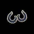 A pair of gold crescent-shaped earrings with deep blue teardrop stones and small clear accent stones, displayed on a dark reflective surface with the brand name 'SAHELI MY BEST FRIEND' above them.