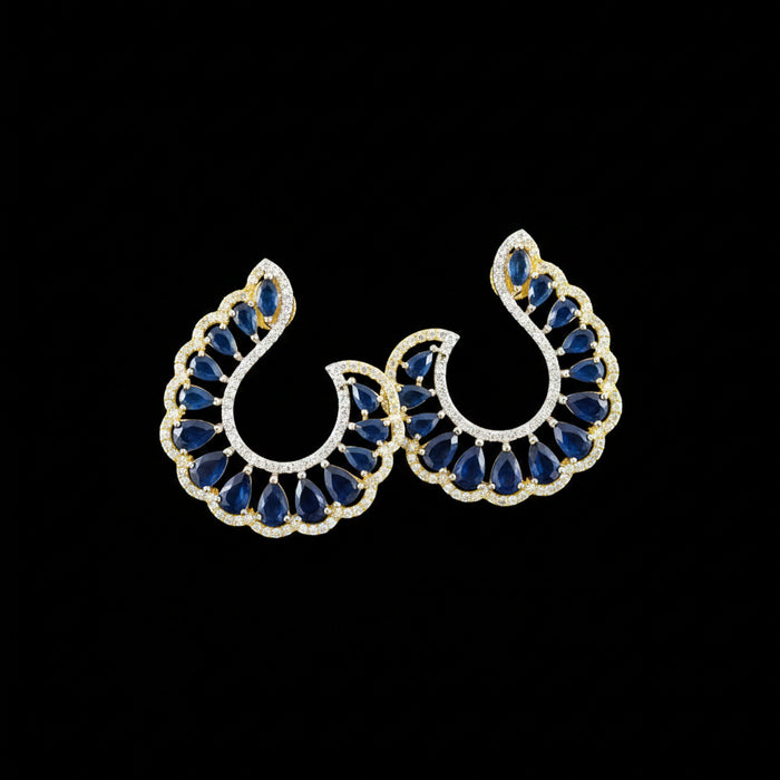 A pair of gold crescent-shaped earrings with deep blue teardrop stones and small clear accent stones, displayed on a dark reflective surface with the brand name 'SAHELI MY BEST FRIEND' above them.
