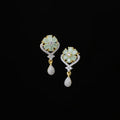 A pair of gold and white stone drop earrings with flower-shaped studs and teardrop charms, displayed on a dark gray velvet jewelry stand against a dark gray background.
