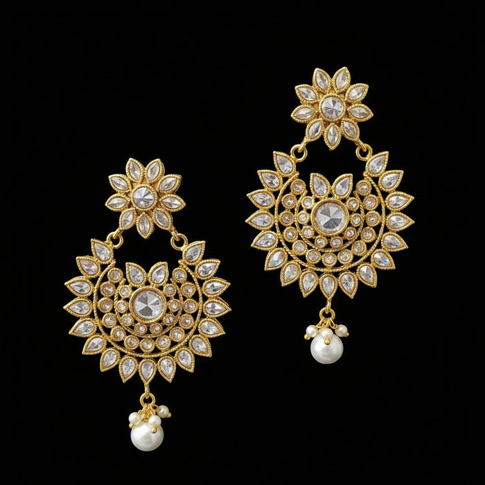 A pair of gold-toned traditional Indian dangle earrings with clear stones and white pearl drops, displayed on a black stand against a gray background with the brand name SAHELI MY BEST FRIEND.