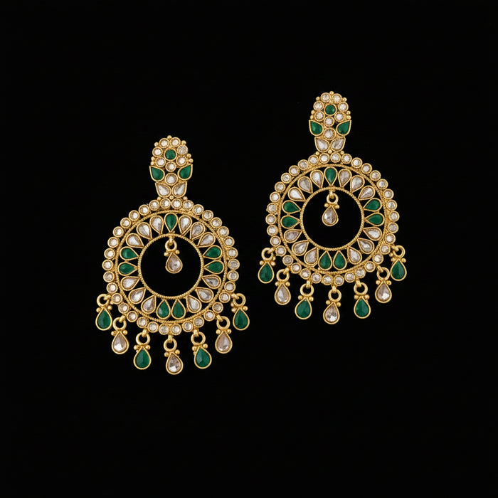 A pair of gold-plated Indian jhumka earrings decorated with green enamel and clear stones, displayed on a dark gray reflective surface with a 'SAHELI MY BEST FRIEND' logo watermark at the top.