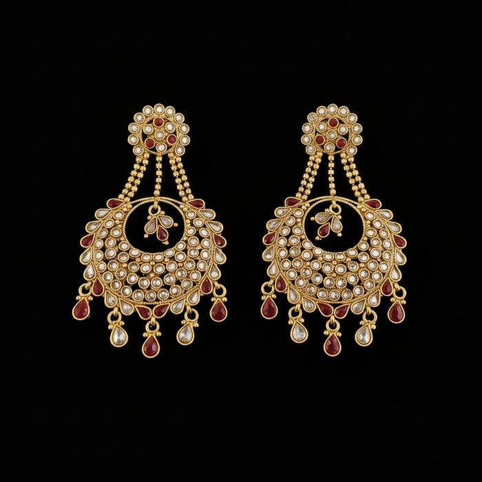 A pair of traditional Indian gold-plated chandelier earrings decorated with red enamel and clear stones, displayed on a dark reflective surface with the Saheli Jewellery brand logo visible in the background.