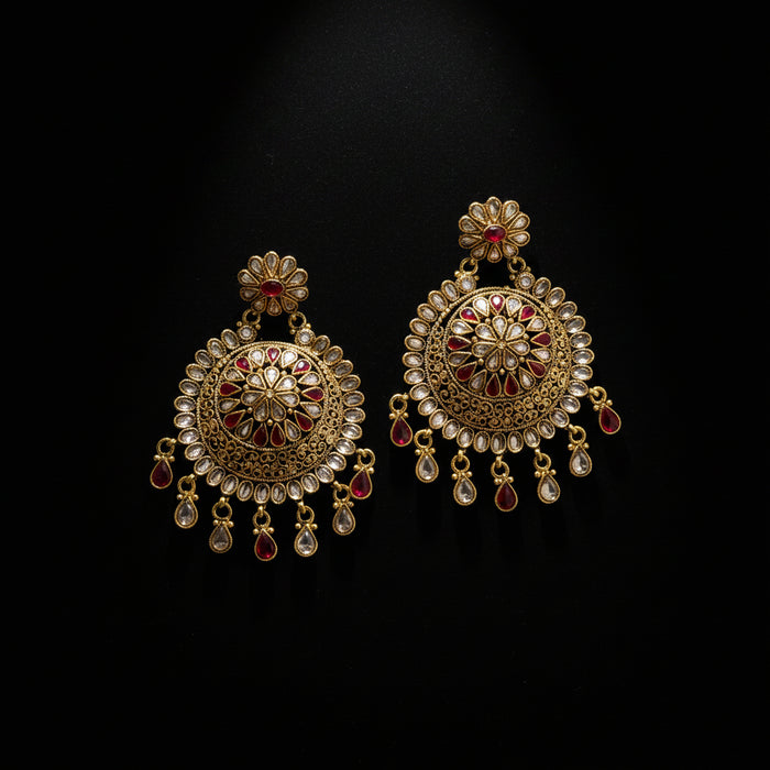 A pair of ornate gold-toned Indian jhumka earrings decorated with red enamel and clear stone accents, displayed on a dark gray surface with a 'SAHELI MY BEST FRIEND' logo overlay.
