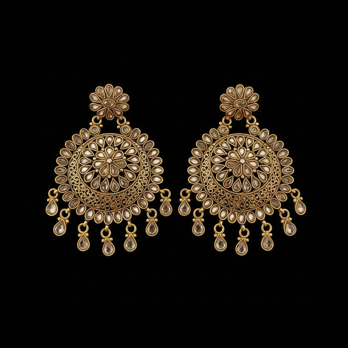 A pair of ornate gold-toned Indian jhumka earrings with intricate filigree work, floral stud tops, and dangling teardrop charms, displayed on a dark gray reflective surface with the Saheli brand logo above.