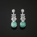 A pair of silver-tone drop earrings featuring a cluster of clear stones in a floral pattern above a teardrop-shaped light green jadeite stone, displayed on a dark reflective surface.