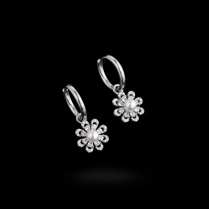 A pair of silver hoop earrings with dangling flower charms, each featuring a central white pearl surrounded by small clear rhinestones, displayed on a white card over a blue background with green leaves in the corner.