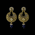 A pair of traditional Indian gold-toned dangle earrings with intricate filigree, white teardrop inlays, small clear stones, and dark blue bead drops, displayed on a dark reflective surface.