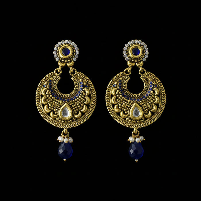 A pair of traditional Indian gold-toned dangle earrings with intricate filigree, white teardrop inlays, small clear stones, and dark blue bead drops, displayed on a dark reflective surface.