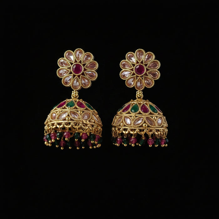 A pair of gold-plated Indian jhumka earrings decorated with clear crystals, pink gemstones, and green gemstones, displayed on a solid dark pink background.