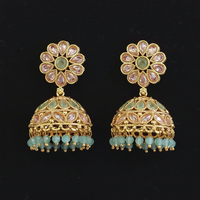 A pair of gold-plated Indian jhumka earrings with light green stone inlays, clear rhinestone flower tops, and dangling light green bead tassels, displayed on a solid dark red background.
