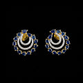 A pair of gold crescent-shaped earrings with blue teardrop stones and clear pavé stones, displayed on a gray reflective surface with the SAHELI MY BEST FRIEND logo above.