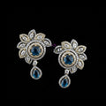A pair of ornate floral drop earrings displayed on a dark gray stand against a gray background, with the brand name 'SAHELI MY BEST FRIEND' printed above. The earrings feature gold and silver-tone metal settings, clear pavé stones, and deep blue teardrop and round center stones.