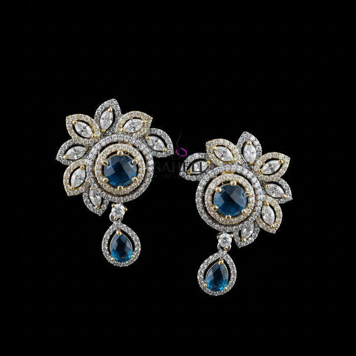 A pair of ornate floral drop earrings displayed on a dark gray stand against a gray background, with the brand name 'SAHELI MY BEST FRIEND' printed above. The earrings feature gold and silver-tone metal settings, clear pavé stones, and deep blue teardrop and round center stones.