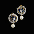 A pair of gold and silver-toned Indian Kundan earrings featuring peacock motifs with deep blue and white stones, finished with a dangling white faux-pearl bead, displayed on a dark reflective surface with the 'SAHELI MY BEST FRIEND' logo in the background.