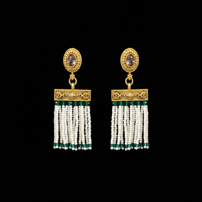 A pair of ornate gold-toned dangle earrings displayed on a dark gray Y-shaped jewelry stand. Each earring features a round gold top with a small oval stone, a rectangular gold connector, and multiple strands of white beads with small green accents.