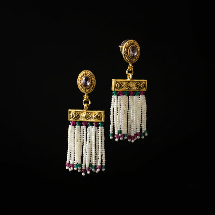 A pair of gold-toned traditional dangle earrings with oval purple stones, ornate gold plates, and white beaded tassels accented with green and pink beads, displayed on a gray jewelry stand.
