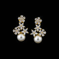 A pair of gold and white pearl drop earrings with clear crystal floral clusters, displayed on a dark gray velvet jewelry stand against a dark gray background.