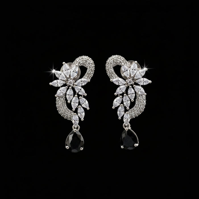 A pair of silver-tone drop earrings with clear marquise and round stones forming a floral heart design, each ending in a black teardrop stone, displayed on a black stand against a gray background. The brand logo 'SAHELI MY BEST FRIEND' is visible in the top left corner.