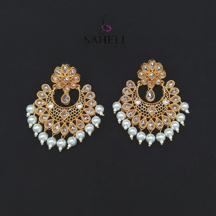 A pair of gold-toned traditional Indian chandbali earrings decorated with white faux pearls and clear stones, displayed on a black background with the SAHeli brand logo above.