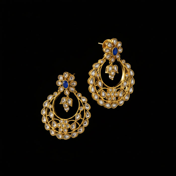 A pair of gold-toned traditional Indian chandbali earrings embellished with white stones and a small blue stone, displayed on a dark gray reflective surface.