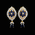 A pair of gold floral drop earrings with deep blue oval stones, surrounded by clear marquise and round crystals, displayed on a black stand against a gray background with the SAHELI MY BEST FRIEND logo at the top.