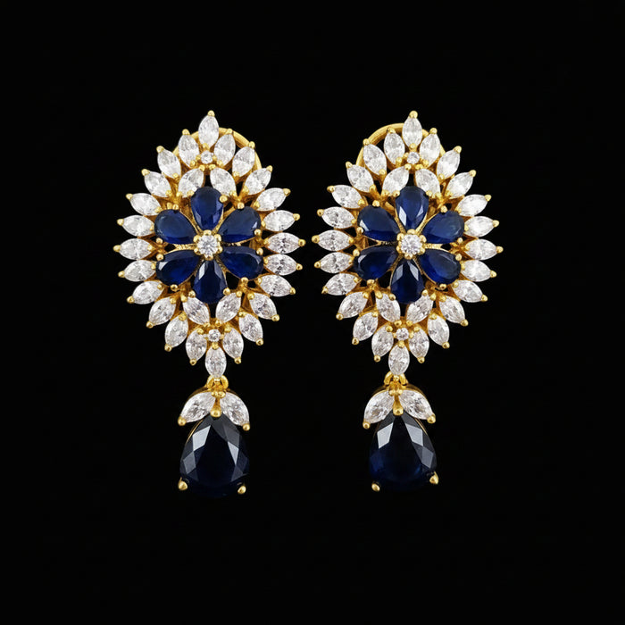 A pair of gold floral drop earrings with deep blue oval stones, surrounded by clear marquise and round crystals, displayed on a black stand against a gray background with the SAHELI MY BEST FRIEND logo at the top.