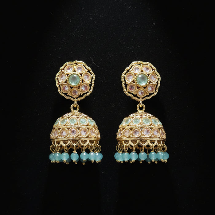 A pair of ornate gold-plated jhumka earrings with green stone accents and light blue bead drops, displayed on a solid dark pink background.