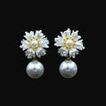 A pair of white flower-shaped drop earrings with pearl centers and gold accents, displayed on a black jewelry stand against a dark background. The brand logo 'SAHELI MY BEST FRIEND' is visible at the top center.